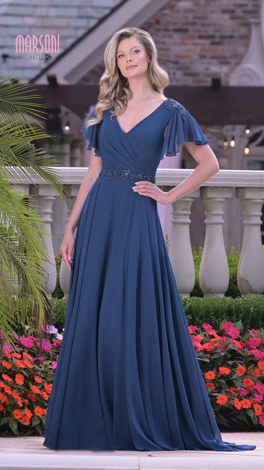 A woman stands outdoors among greenery and flowers, wearing the Marsoni M328 long formal dress with chiffon fabric, flutter sleeves, an a-line skirt, floral details, and a beaded waistband.